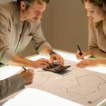 Quelle stratégie immobilière choisir : acheter maintenant ou attendre 2027 ? Un homme et une femme examinent un budget immobilier avec une calculatrice, des plans et une carte non légendée de la région Provence-Alpes-Côte d’Azur sur une table lumineuse.